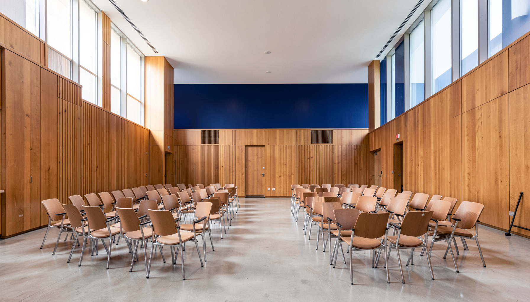 A wide-angle view of a modern community meeting space with warm wood paneling, rows of chairs arranged in neat arcs, and soft natural light streaming through tall windows. The room feels open, civic, and welcoming.